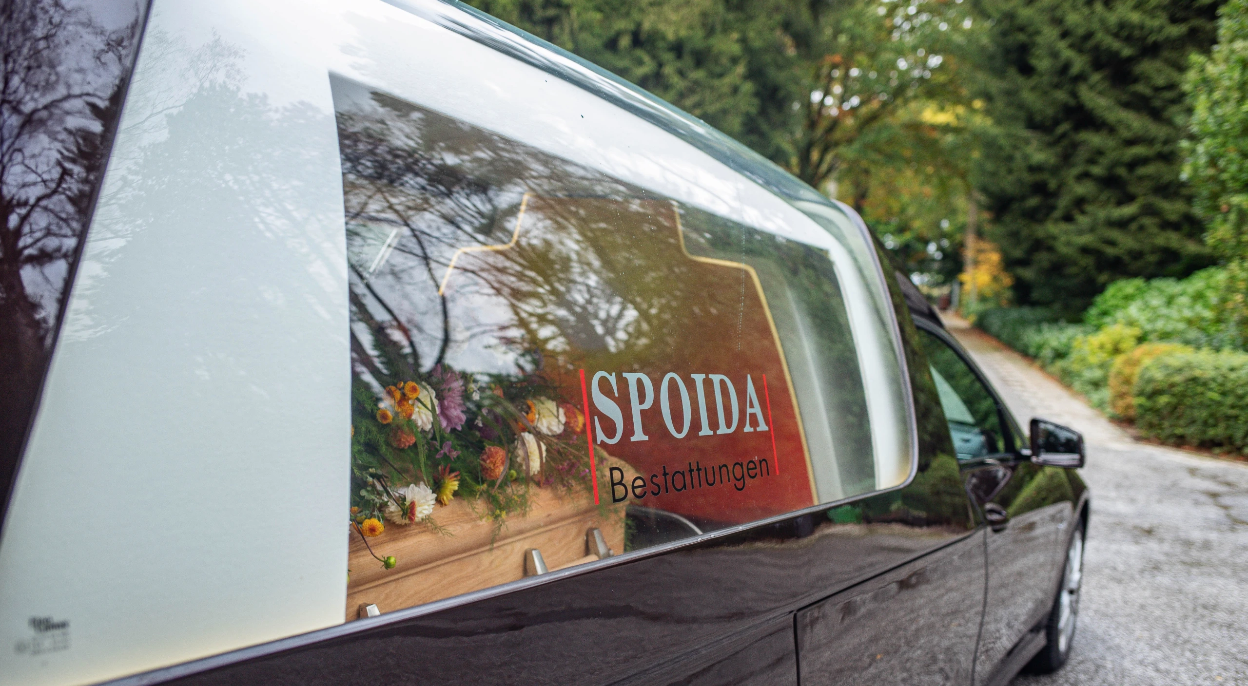 Bestattungswagen seitlich im Anschnitt. Fenster mit Logo beschriftet. Blick auf Sarg mit Blumendekoration.