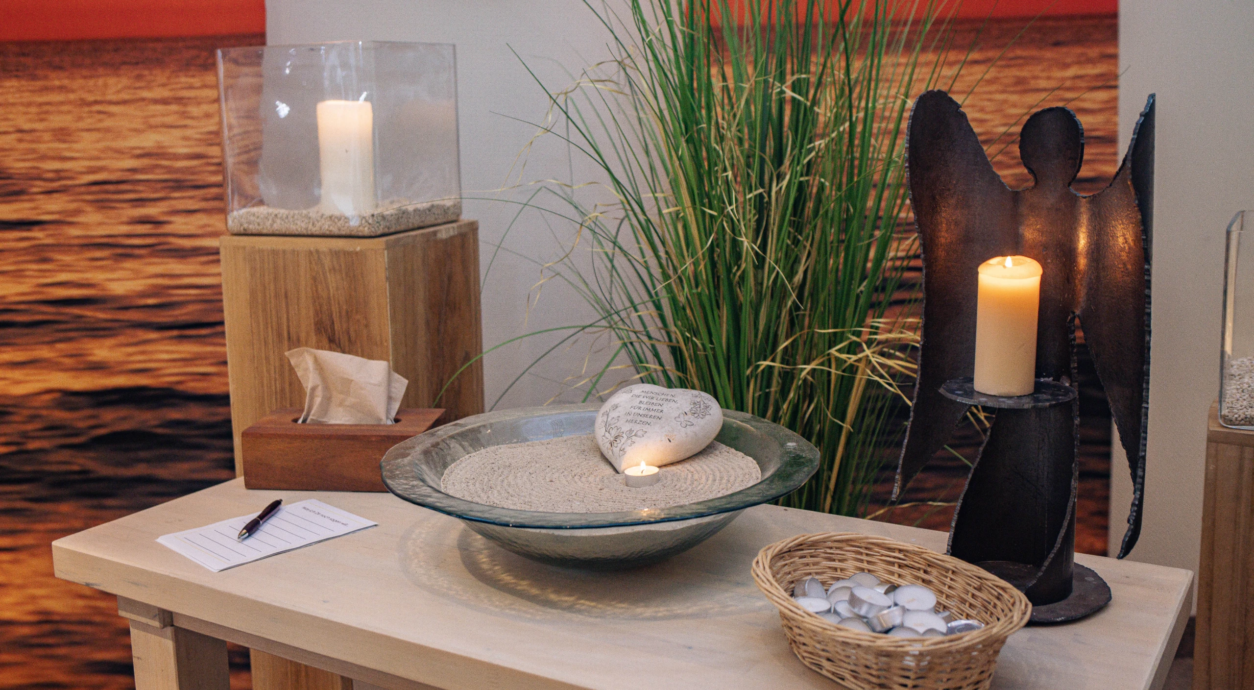 Glasschale mit Sand, Kerze und Steinherz auf Tisch, Kerzenständer in Engelform, Korb mit Teelichtern und Zettel mit Stift