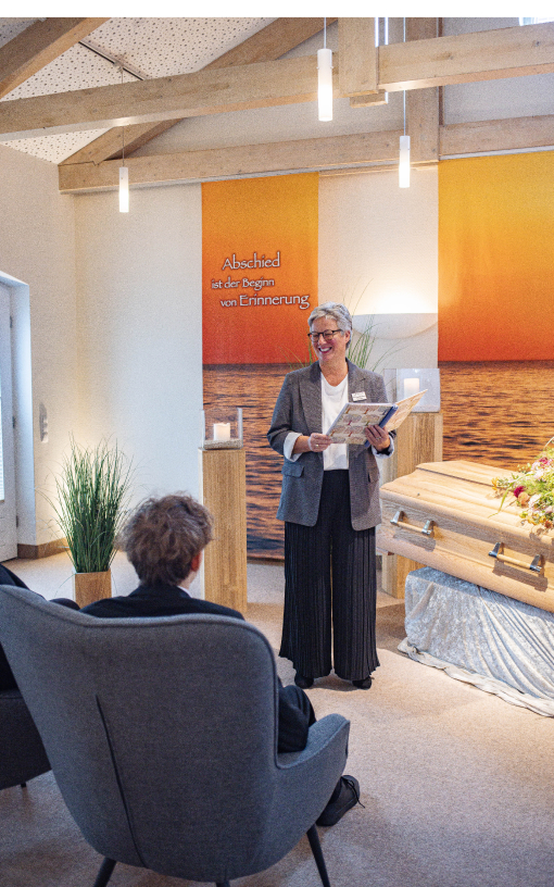 Trauerfeier mit Sarg, Gesteck, Kerzenstehlen und Kunstgras. Mitarbeiterin spricht lächelnd zu sitzenden Angehörigen.