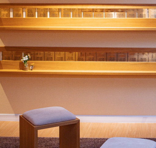 : Zwei Holzregale mit gravierten Schildern der Verstorbenen. Vor einem Schild kleine Blumenvase. Hocker vor Wand