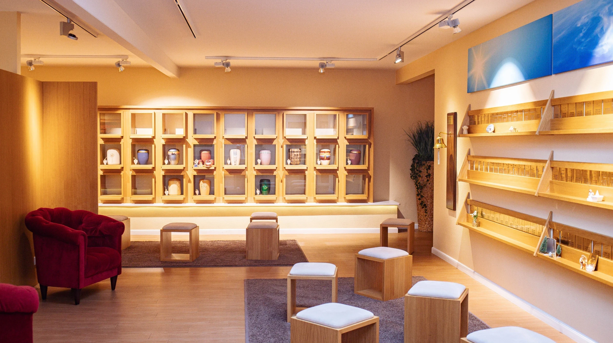 Blick in den beleuchteten Abschiedsraum mit Kolumbarium und Gedenkwand. Sitzhocker und zwei rote Sessel.