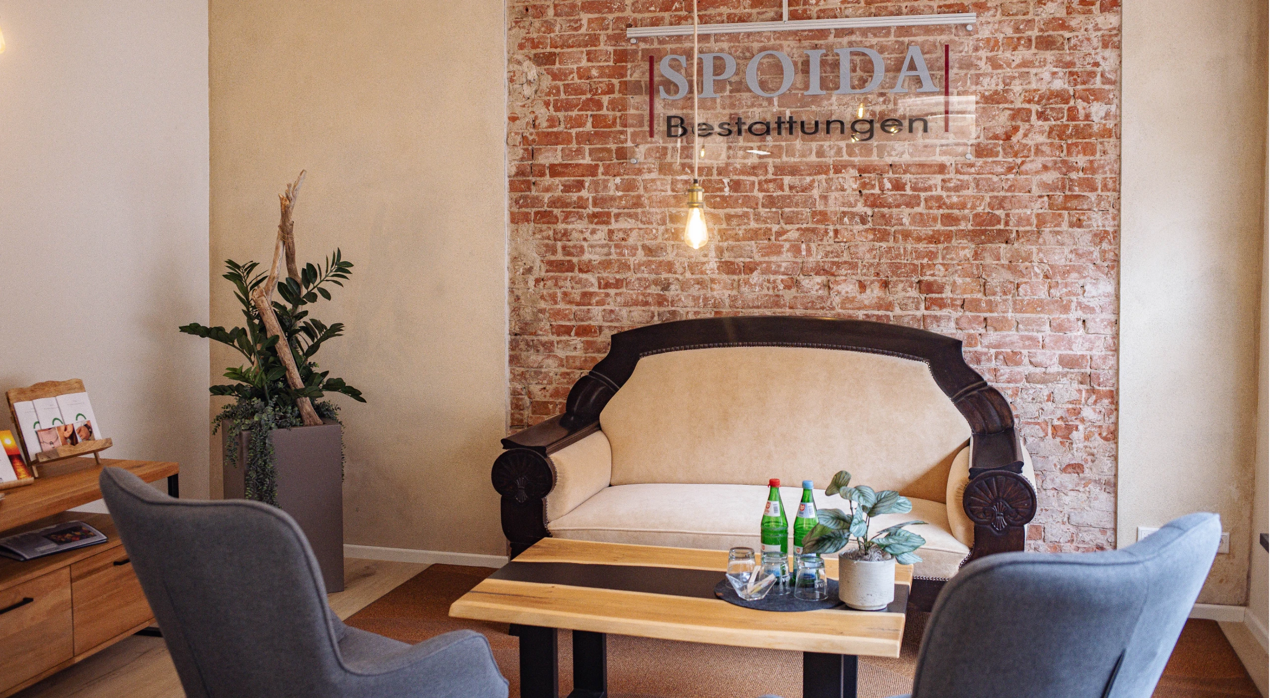 Blick auf Sitzecke mit Sofa, Logo und zwei Sessel. Tisch mit Pflanze, zwei Wasserflaschen und Gläsern, Regal an der Seite