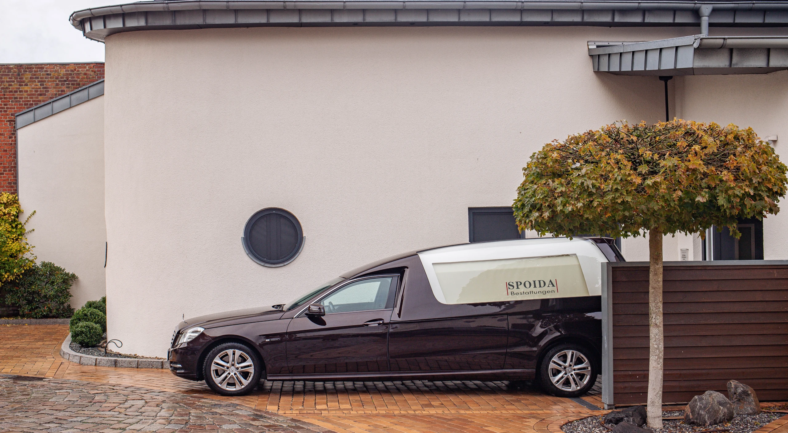Foto: Schwarzer Bestattungswagen rückwärts parkend vor Gebäude. Logo hinten, auf weißem Fenster, Baum im Vordergrund.