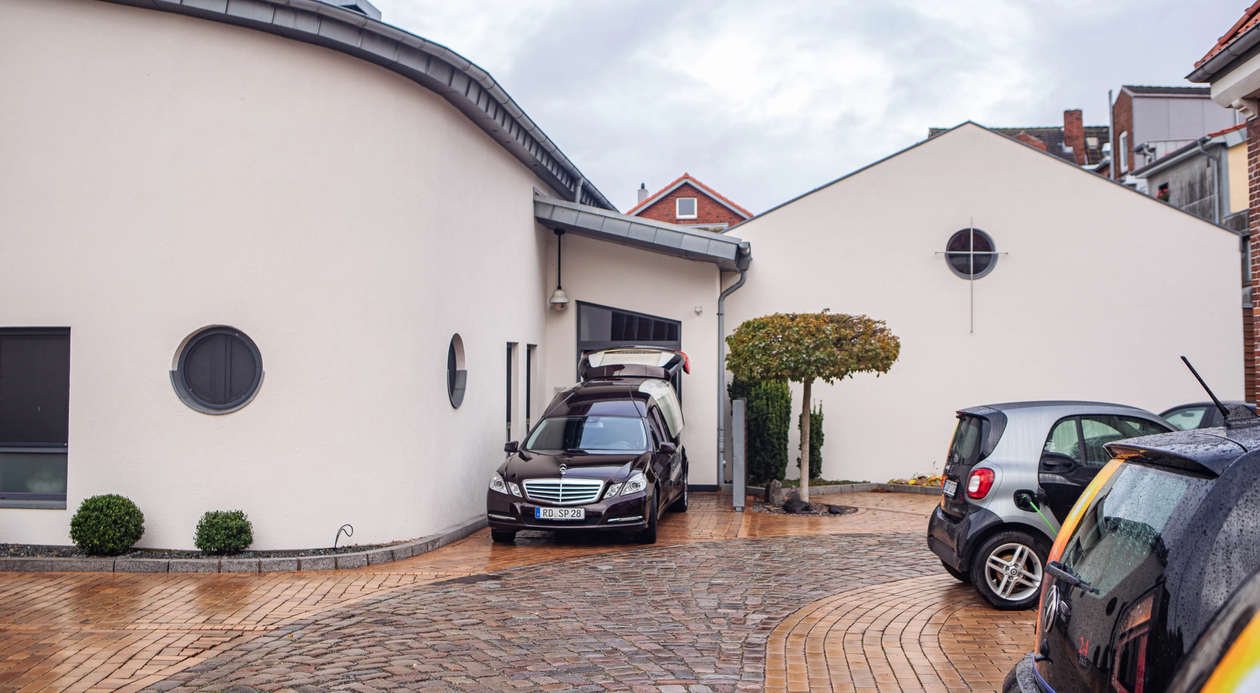 Schwarzer Bestattungswagen rückwärts parkend vor Gebäude von vorne fotografiert, Kofferraum geöffnet. Rechts weiterer Fuhrpark