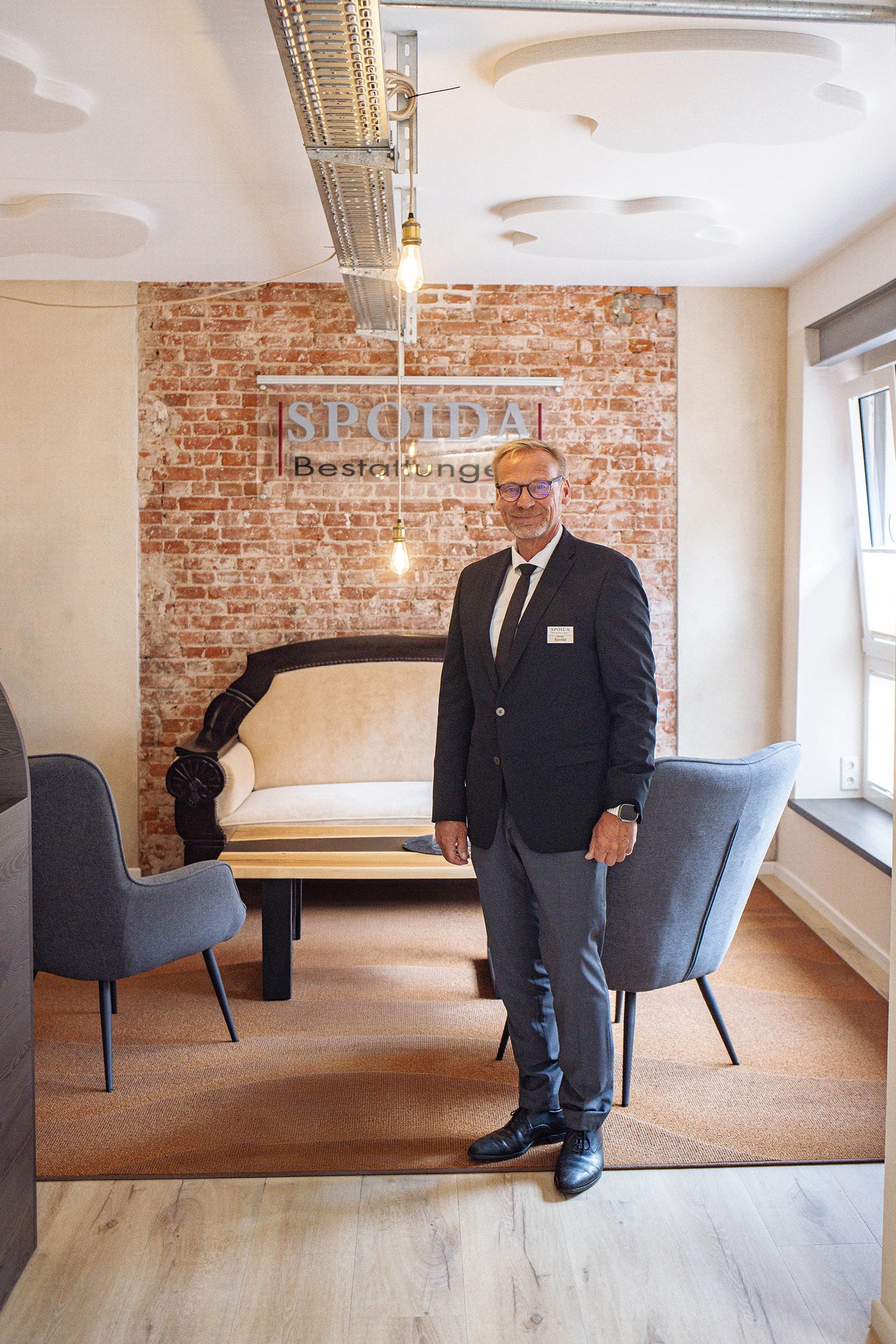 Georg Spoida mit Jackett und Krawatte lächelnd stehend im Eingangsbereich. Sitzecke und Logo im Hintergrund